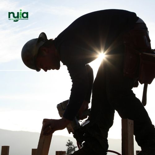 A construction worker wearing a hard hat uses a nail gun on a wooden structure, with the sun shining behind him—a reminder of the importance of seeing a Workers' Comp Doctor in Selden after any job site injury.