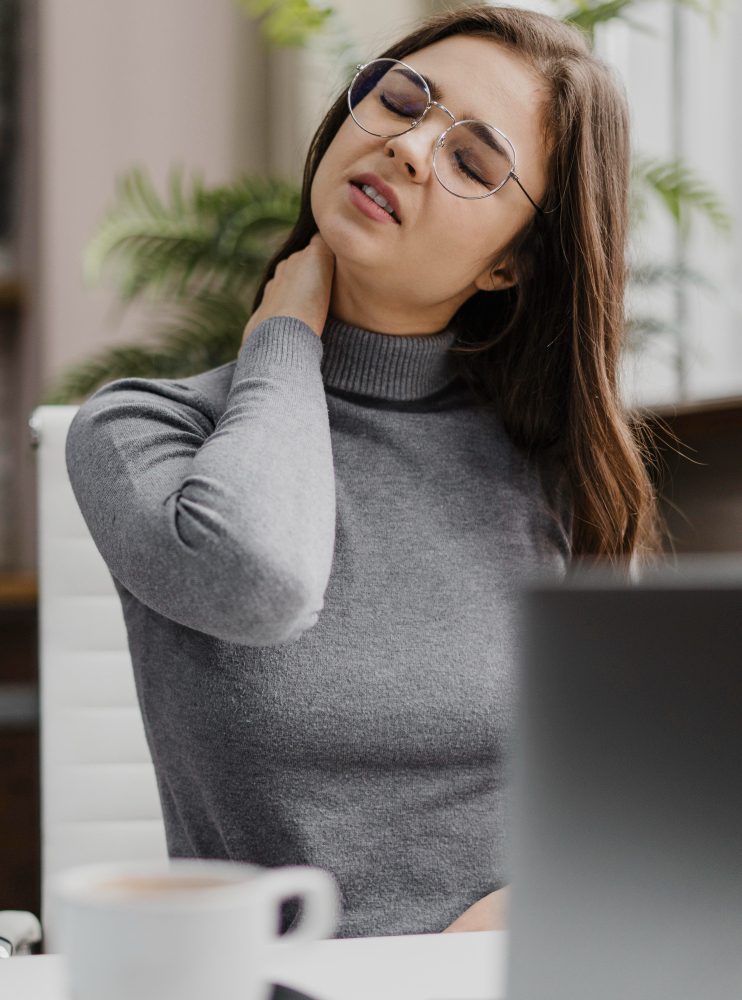 A woman wearing glasses sits at a desk and touches her neck, appearing to feel discomfort. A laptop and coffee cup are in the foreground.