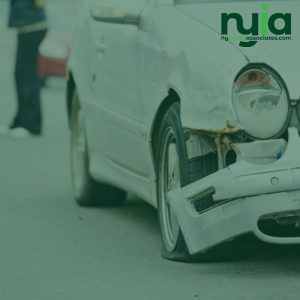 Front side of a white car with visible damage to the bumper and wheel; a person stands blurred in the background. Logo for "ny injury associates" in the top right corner, highlighting no hay culpa doctor en Middle Village services.