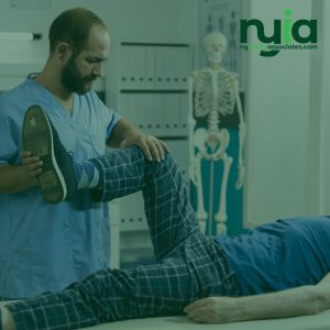 A physical therapist in scrubs bends a patient's knee during an examination in a clinic, with an anatomical skeleton model in the background.