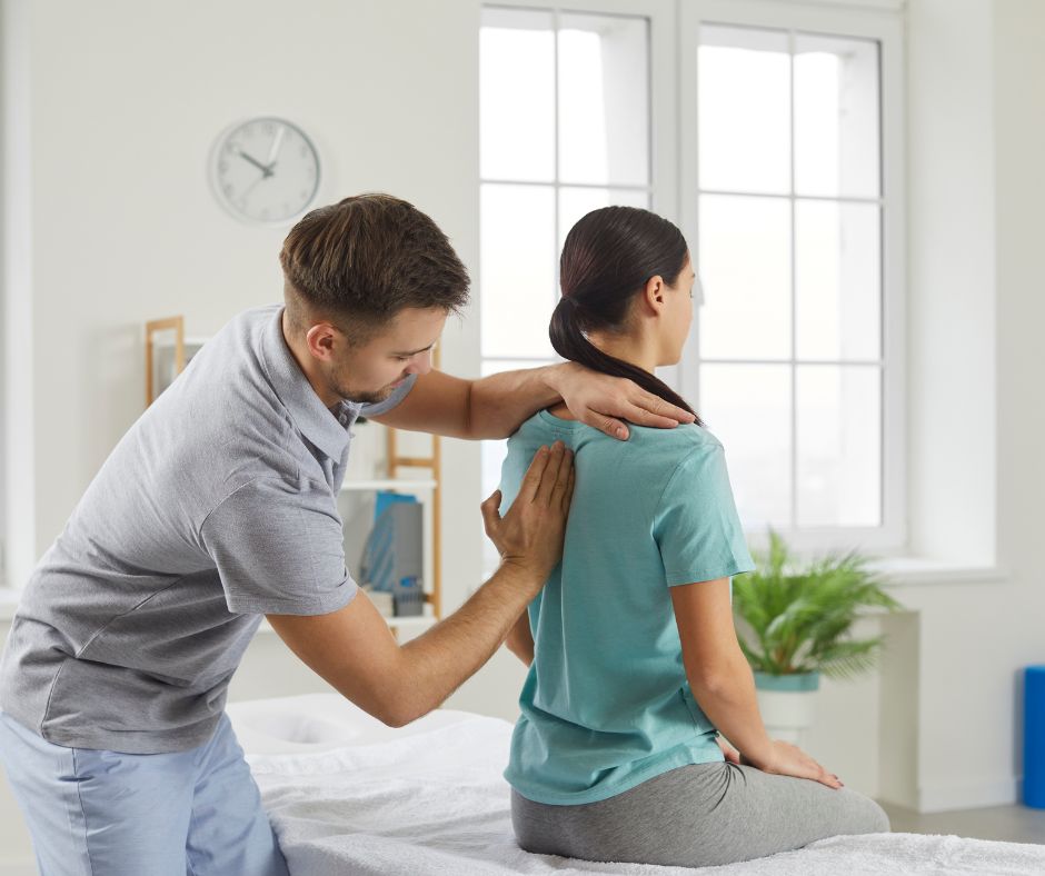 A physical therapist examines a woman’s upper back as she sits on an exam table in a bright room with large windows 