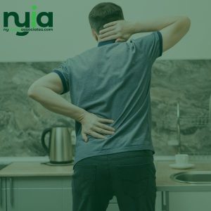 A man standing in a kitchen holds his lower back and neck, appearing to experience discomfort or pain. The logo "nyia associates.com" is visible in the top left corner, highlighting no fault insurance doctors in Lindenwood.